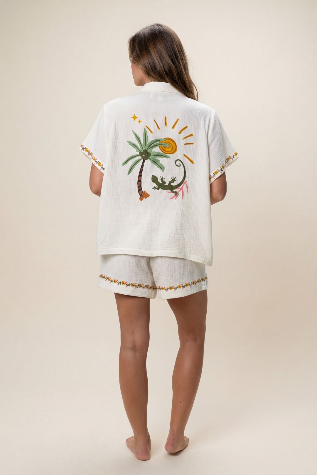 Woman wearing a white embroidered shirt and shorts set with a palm tree design on a beige background